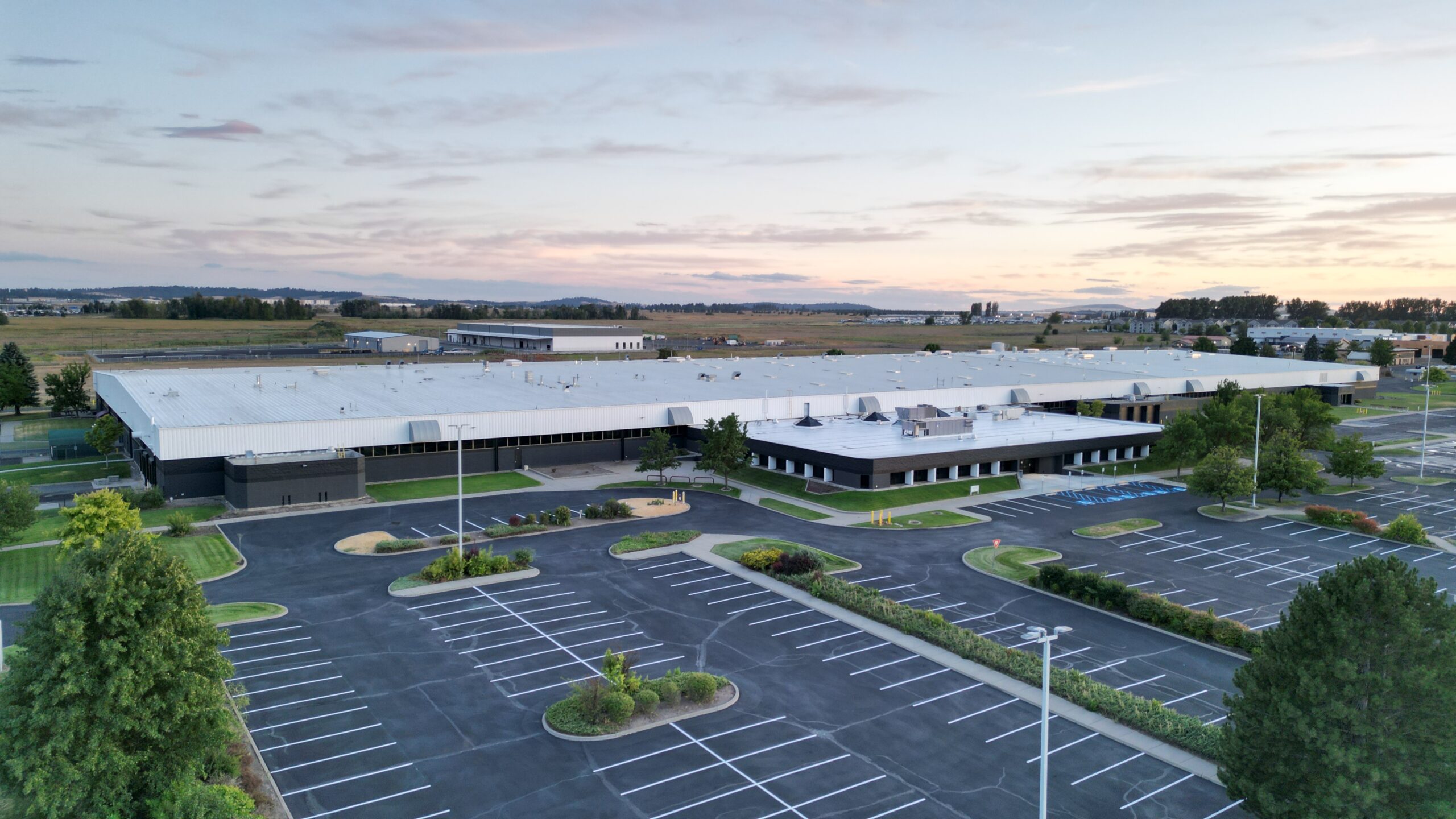 The proposed site of the American Aerospace Materials Manufacturing Center is pictured: a large warehouse at sunset with a parking lot and cars.